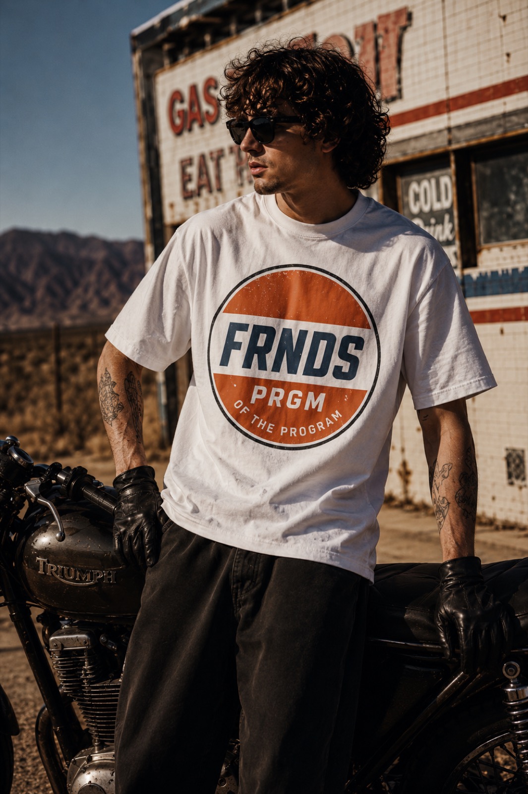 Model wearing the white FRNDS PRGM Moto Tee beside a motorcycle in the desert